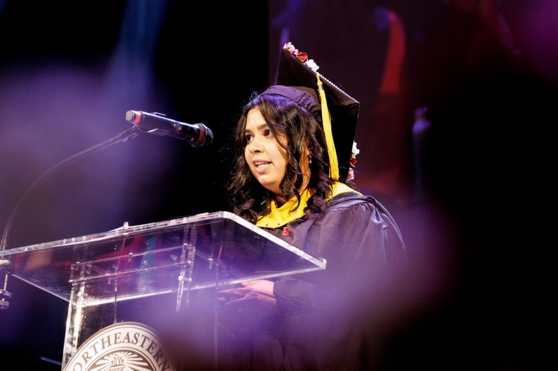 A female graduate wearing a mortarboard and gown speaks at a clear podium with a microphone, illuminated by colorful stage lighting in purple and blue hues.