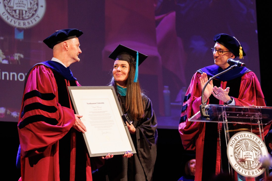 A dean in red and black academic robes presents a framed certificate to a female graduate in a black cap and gown, while another official in purple robes applauds nearby.