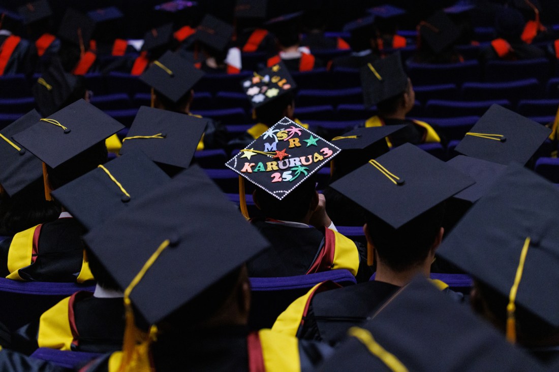 An overhead view of numerous black graduation caps with yellow tassels, with one cap decorated with "KARURU43" text and colorful designs.