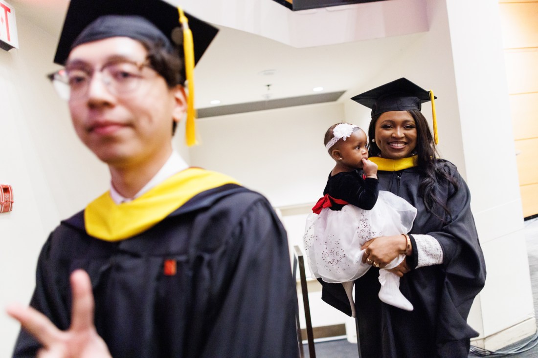 A graduate in a cap and gown walks past while a smiling woman in academic regalia holds a young child during Northeastern University Vancouver Campus's Convocation 2025 in Vancouver, B.C.