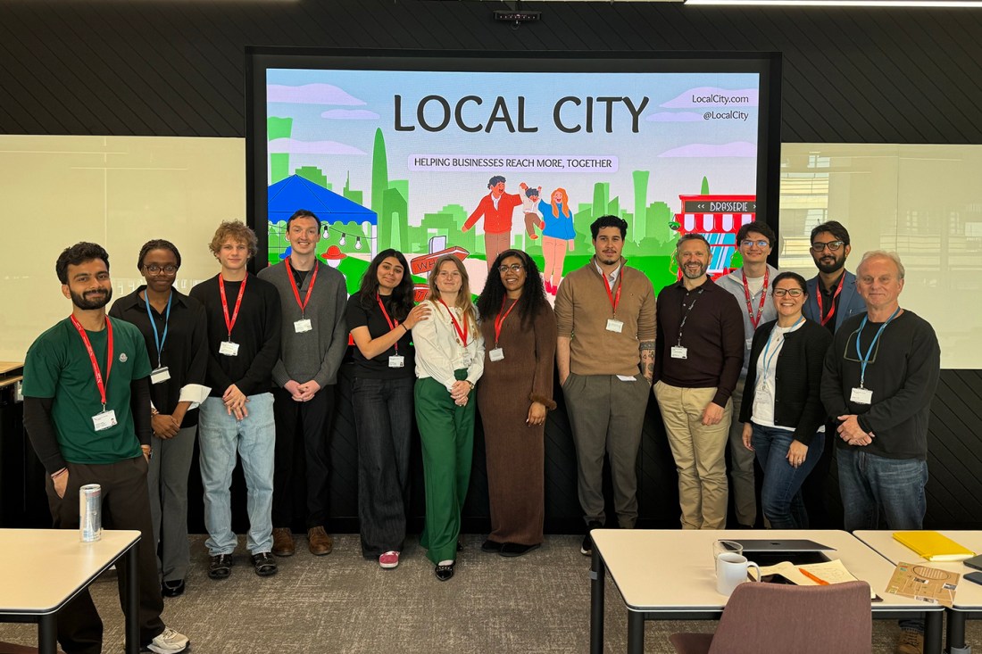 A group of 13 people, diverse in age, gender, and ethnicity, pose together and smile in front of a large screen that reads “LOCAL CITY – Helping Businesses Reach More, Together.” The screen features cartoon-style graphics of people and city landmarks.