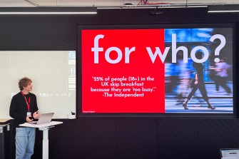 A person wearing jeans, a black sweater, and a red lanyard stands next to a screen presenting a slide that says "for who? '25% of people (18+) in the UK skip breakfast because they are too busy' - The Independent".