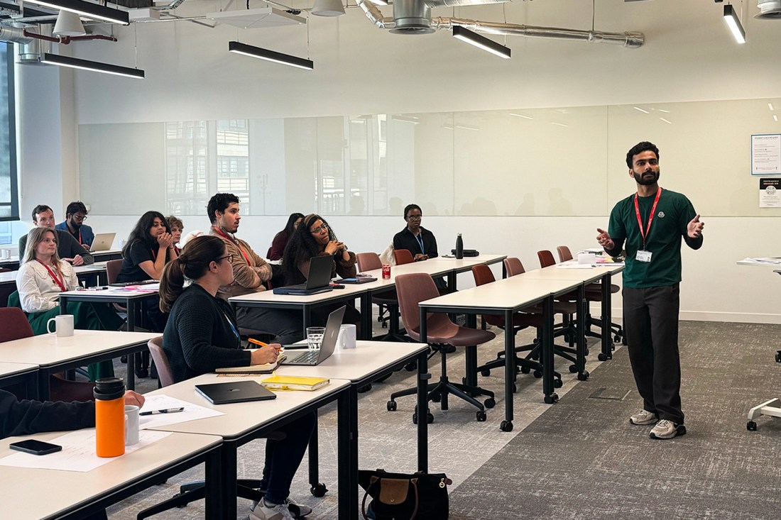 A man wearing a green sweater and red lanyard stands at the front of a classroom speaking to a group of students seated at long desks with laptops and notebooks.