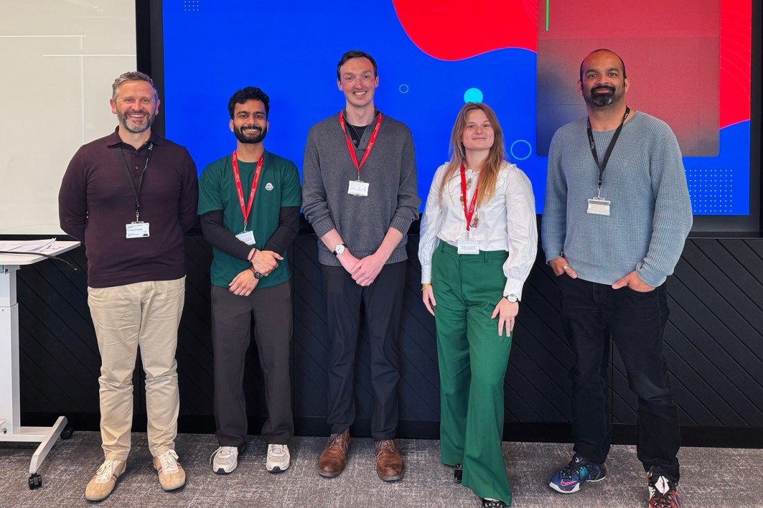 Five people wearing conference badges stand in a row and smile for the camera in front of a presentation screen. 