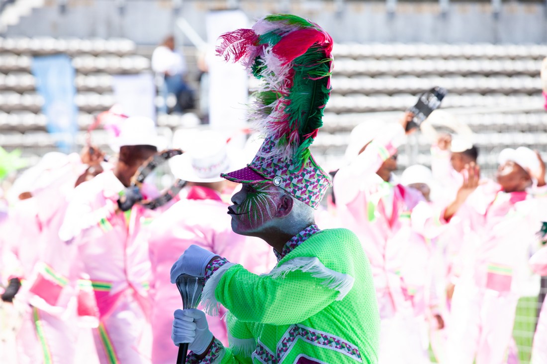 A person wearing a bedazzled green shirt and a hat with a green and pink feather on top dancing in front of a group of people wearing pink matching outfits.