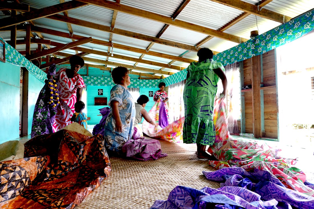 Women and a child gather in a brightly colored room in Fiji, folding and arranging patterned fabrics on a woven floor mat.