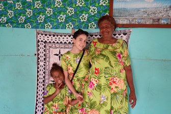 Three people in matching floral dresses pose in front of a turquoise wall, smiling inside a Fijian home.