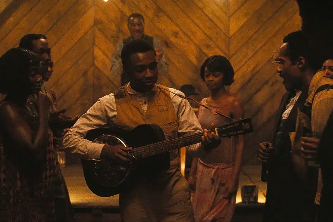 Musician performs on a guitar while people gather around in a dimly lit room.