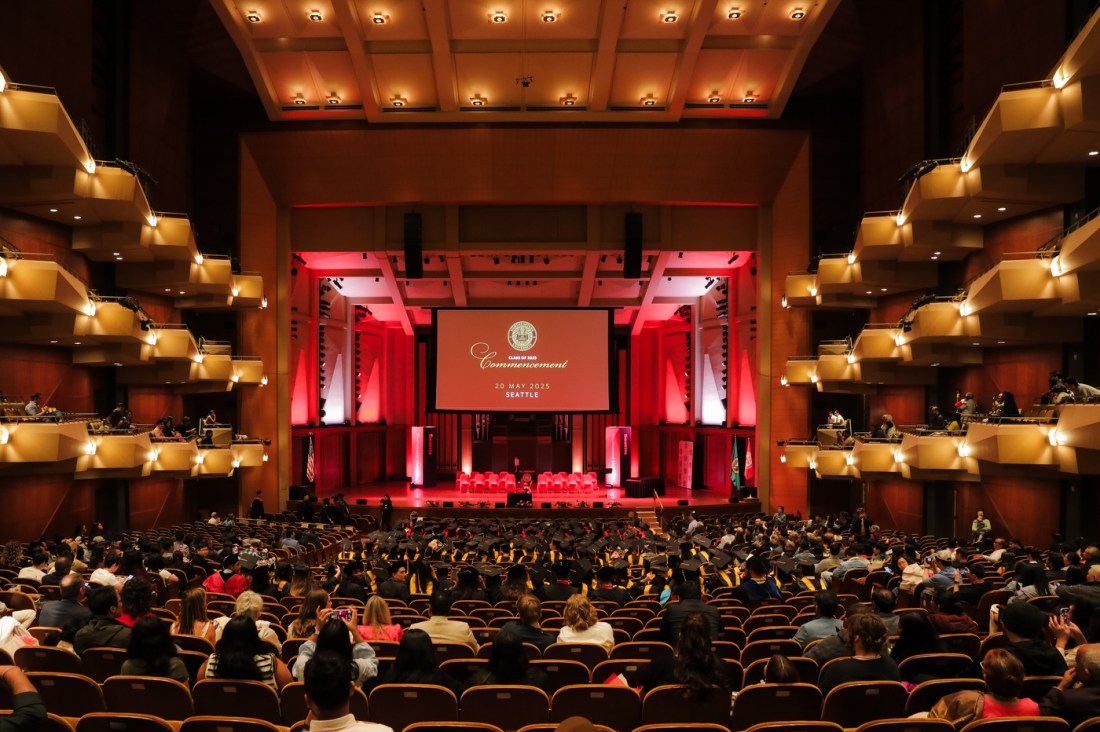 Wide view of the Seattle Campus Commencement Ceremony at Benaroya Hall, showing a packed auditorium with red lighting on the stage and a commencement banner.