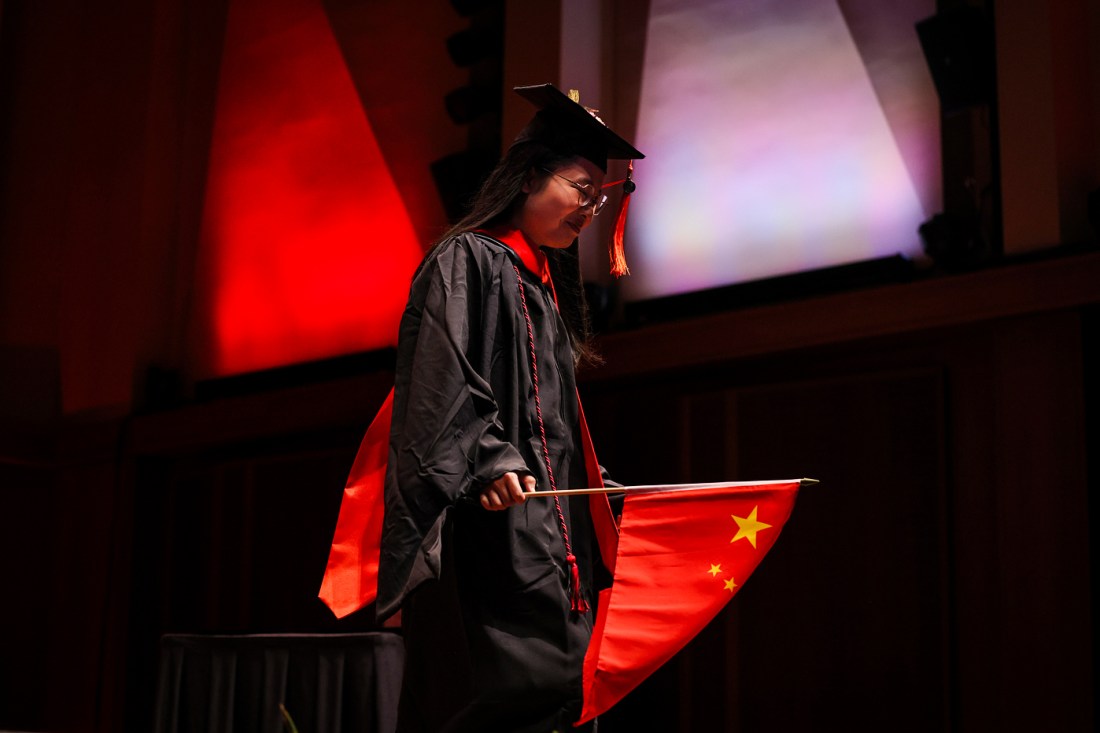 A graduate in a cap and gown walks across the stage holding a Chinese flag during the Seattle Campus Commencement Ceremony at Benaroya Hall.
