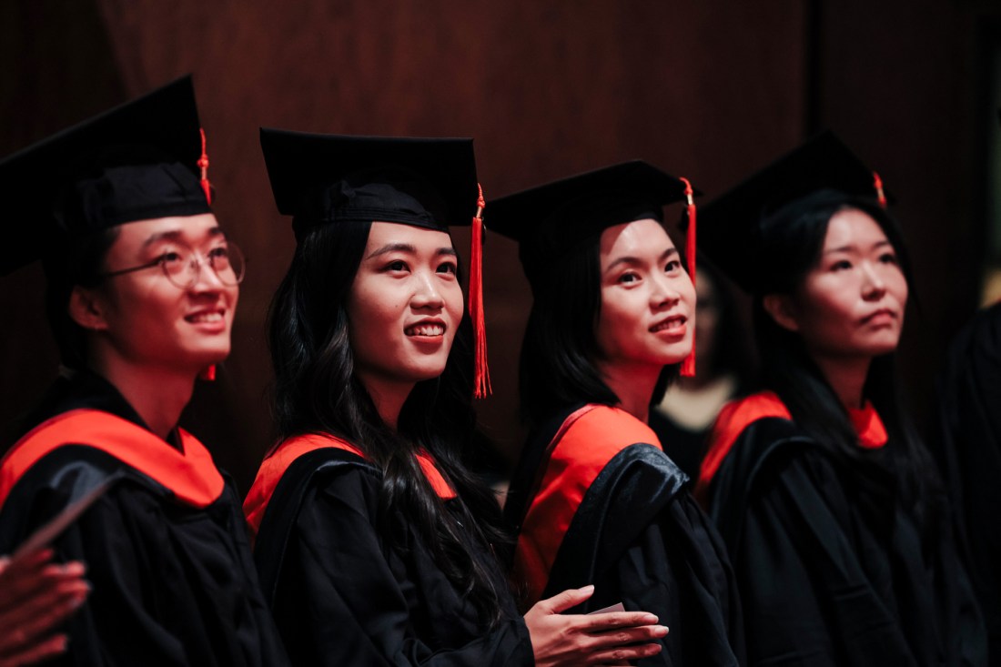 A row of four Northeastern graduates in black caps and gowns with red accents smile and look toward the stage during the Seattle Commencement ceremony.