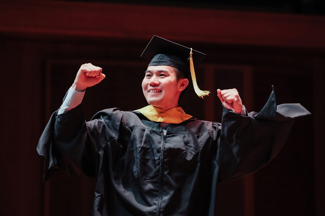 A graduate in a bblack cap and gown with yellow accents raises both fists in celebration while walking across the stage.