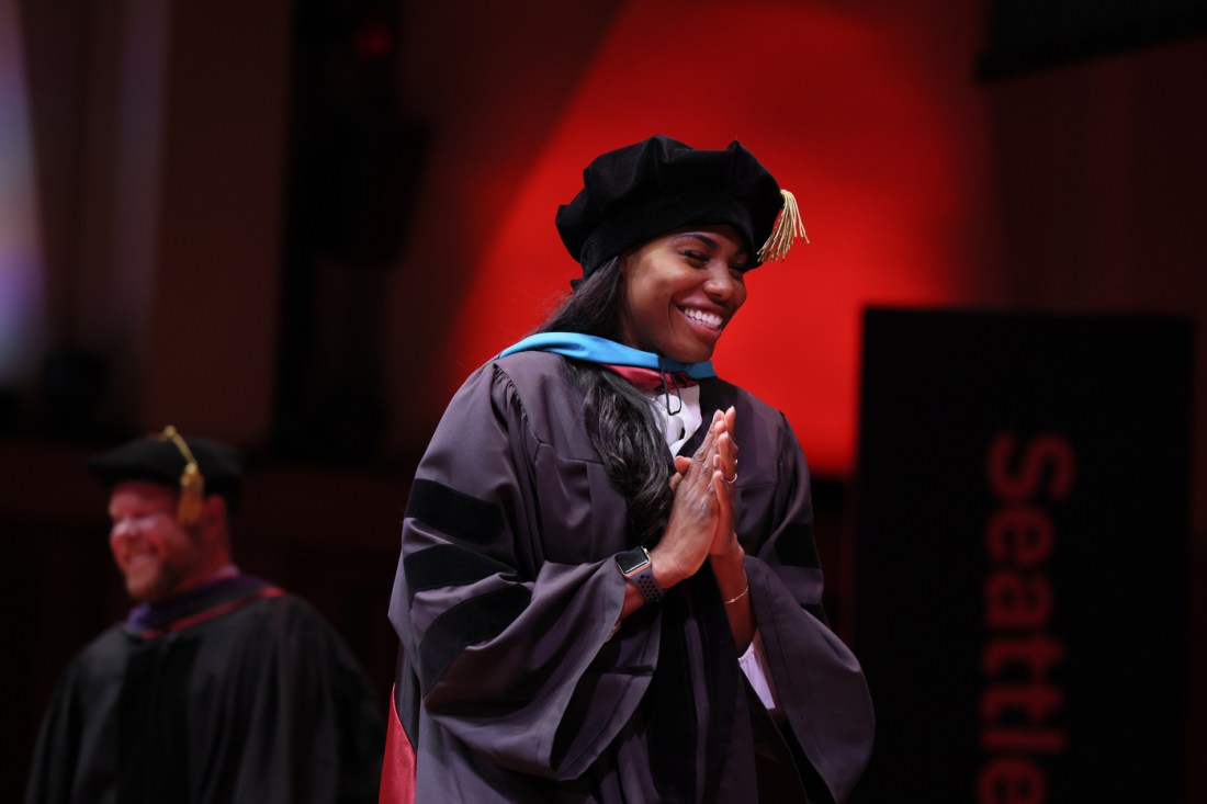 A graduate in a doctoral cap and gown stands on stage smiling with hands clasped, as red light glows in the background.