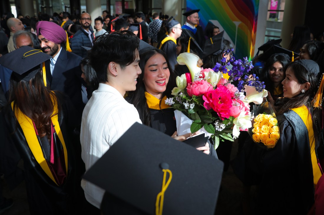 A graduate receives a large bouquet of flowers from another person while standing amongst a crowd of other graduates at the Seattle Campus Commencement Ceremony.