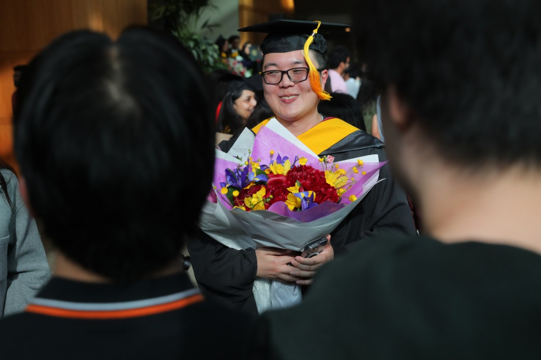 A smiling graduate wearing a black cap and gown with gold accents poses with a bouquet of flowers. 