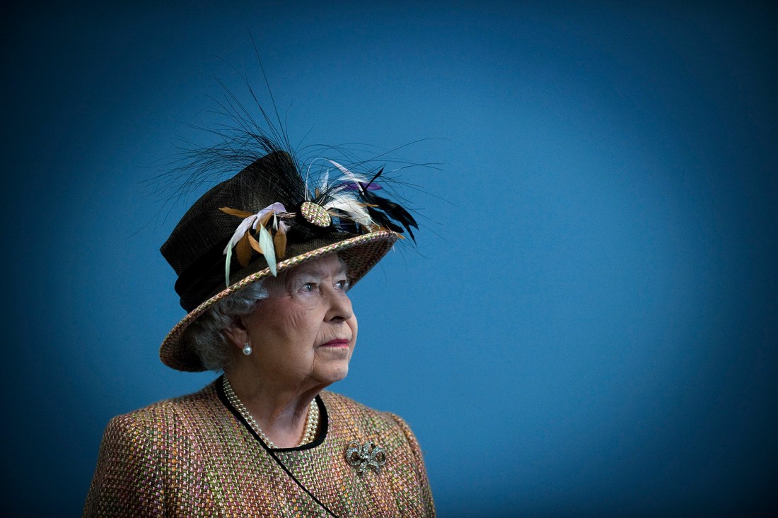 Queen Elizabeth wearing a black hat with feathers on it posing for a portrait on a blue background.