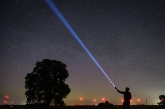 Silhouette of a person pointing a flashlight into a star-filled night sky beside a large tree.