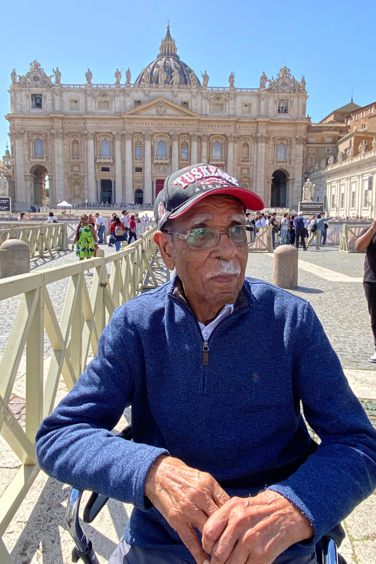 Daniel Keel wearing his TUSKEGEE hat sits in front of an ornate building in Vatican City.