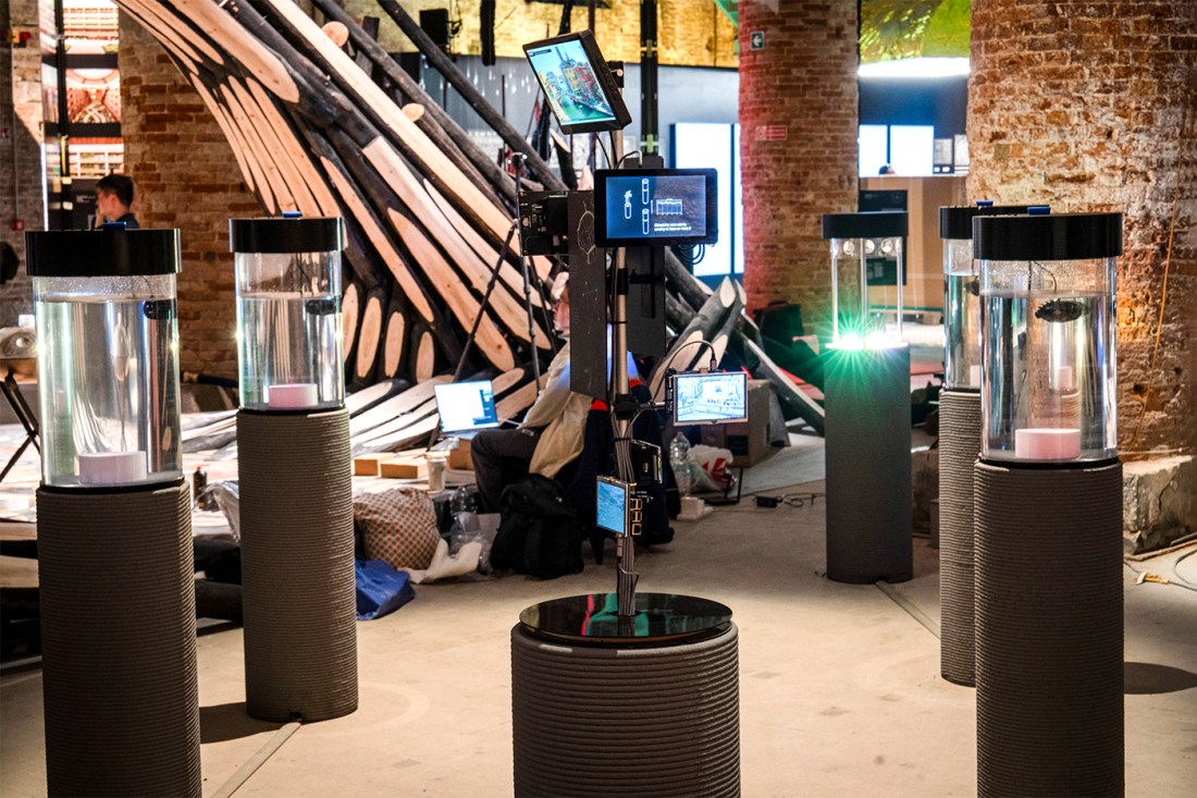 Indoor installation featuring cylindrical water-filled tanks on pedestals arranged in a circle around a central pole with multiple monitors displaying digital interfaces. The background includes a large wooden architectural structure and people working on laptops in a rustic, brick-walled space.