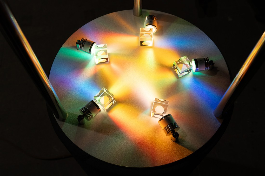 Close-up of a circular table with six prism-like glass structures mounted on lightbulbs, projecting colorful, overlapping rainbows of light onto the surface.