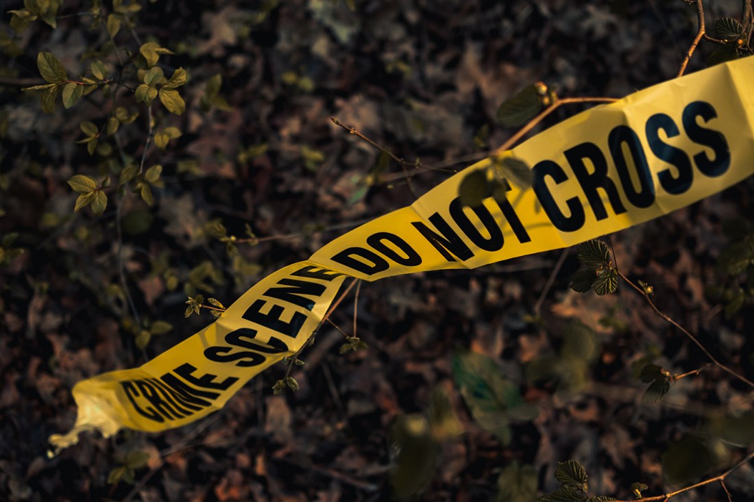 A section of yellow crime scene tape reading “CRIME SCENE DO NOT CROSS” stretches across a wooded area with leaves and branches in the background.