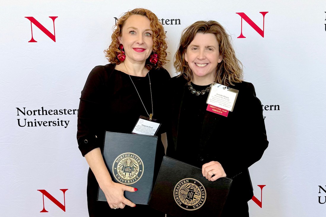 Jennifer Kirwin and Margarita DiVall standing in front of a Northeastern branded backdrop posing with certificates in Northeastern branded folders. 