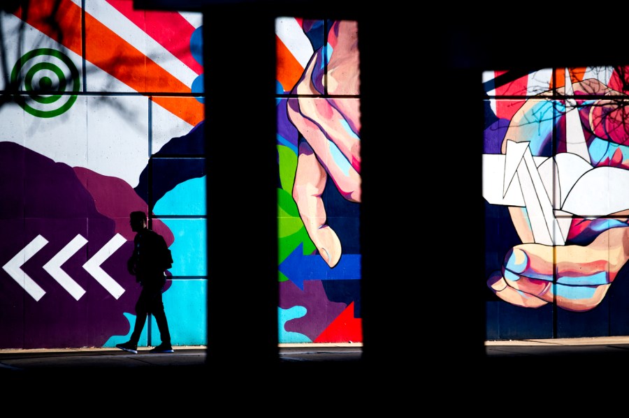 Pieces of two murals, separated by columns. Both have bright vivid colors and stripes, with other graphical elements such as arrows. On the right, a hand holding an origami crane draws focus.