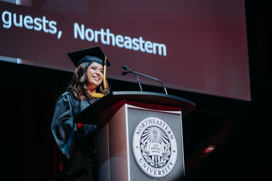 A person wearing black commencement regalia with yellow accents stands at a podium in Meridian Hall smiling.