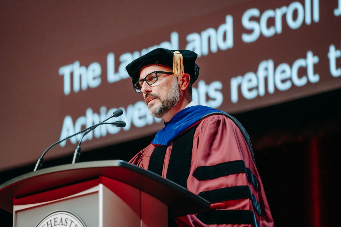 A faculty member wearing maroon regalia with black accents and a blue hood stands at a podium. 