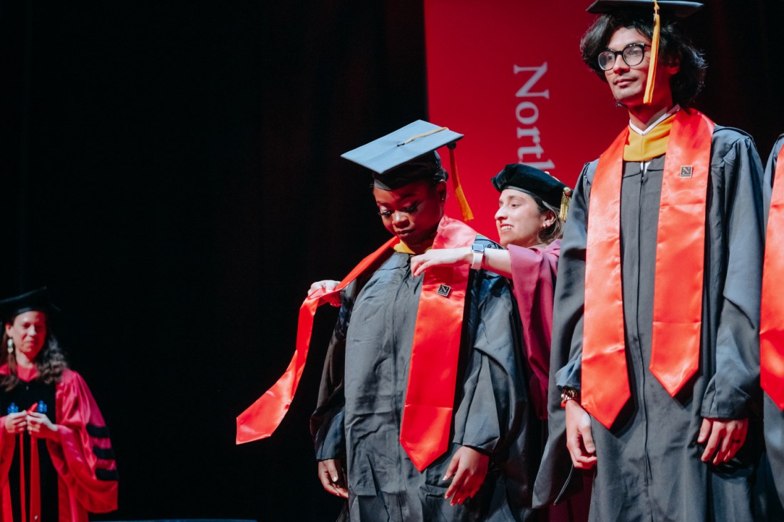 A faculty member helps adjust the red sash of a graduate's regalia from behind. Another graduate wearing the same regalia stands next to them. 