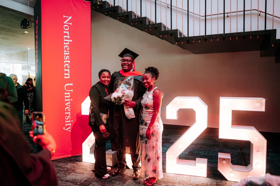 A graduate wearing black commencement regalia with red accents poses in front of large light up numbers that write out '2025'. 