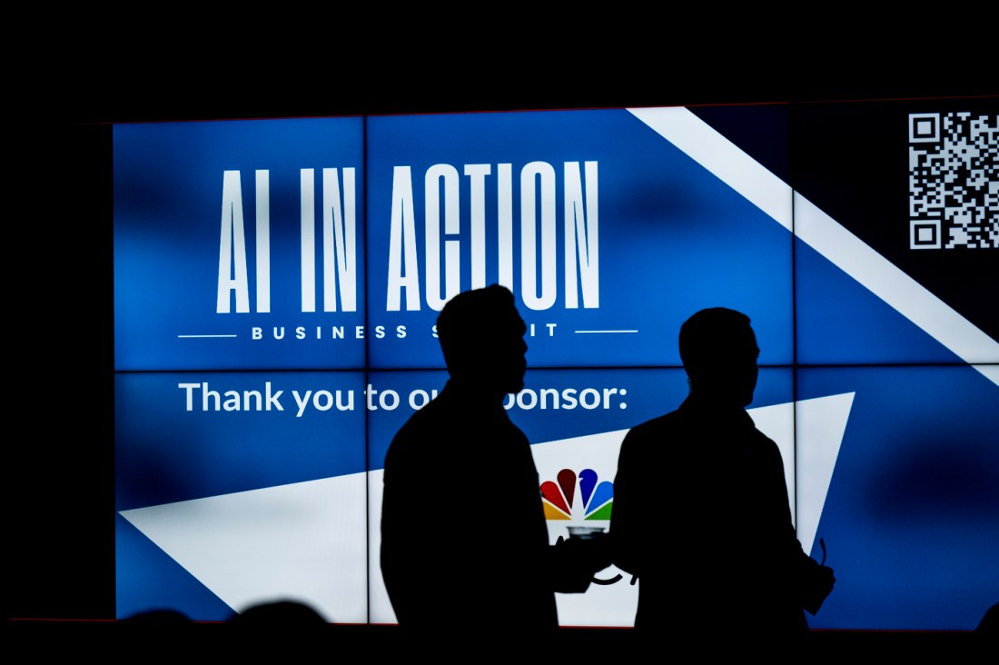 The silhouette of two people standing in front of a blue screen that says 'AI IN ACTION' in large white letters on a blue background. They are at the AI in Action Business Summit.