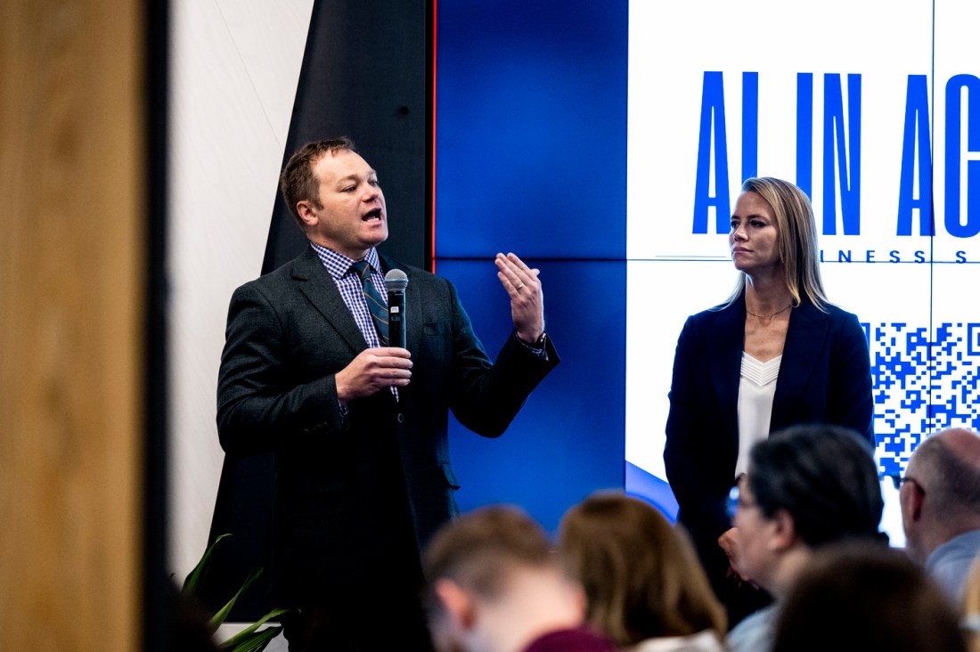 Two people standing together on stage at the AI in Action Business Summit - a man in a dark suit speaking into a microphone and a woman in professional attire, both positioned in front of a conference backdrop.