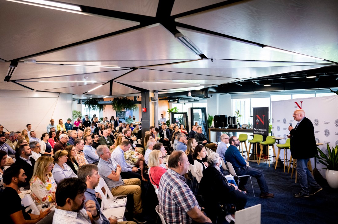 Usama Fayyad stands at the front of the room while speaking during the AI in Action Business Summit. He is standing in front of a crowd of people seated, listening to him. 