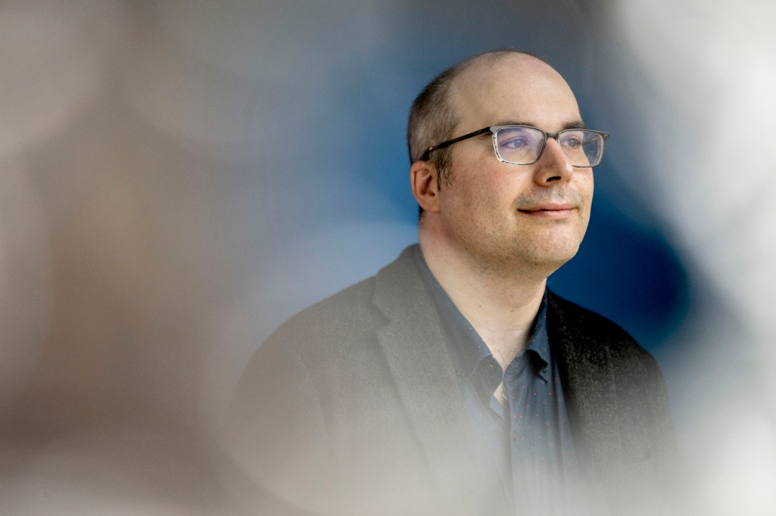 A professional headshot of Michael Correll wearing glasses and a blue collared shirt under a jacket, photographed against a softly blurred blue gray background.