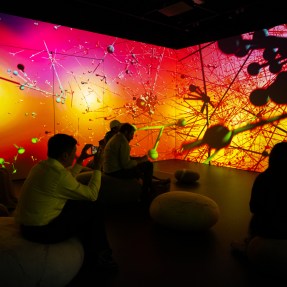 People sit in a dark room surrounded by colorful, abstract visuals projected on the walls.