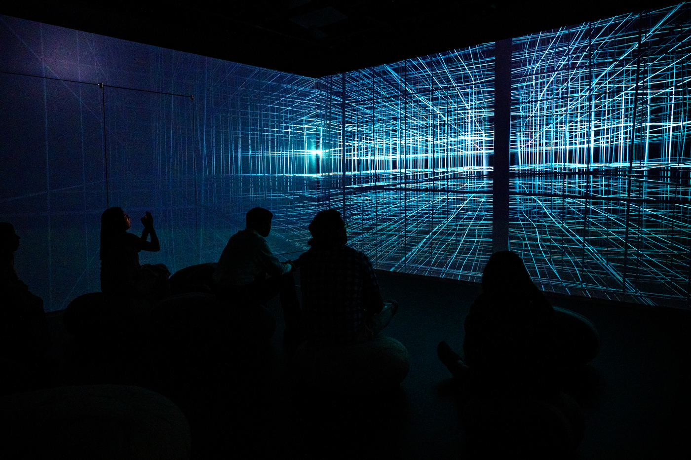 People sit in a dark room watching a glowing digital grid projection at the Data | Art Symposium immersive installation.