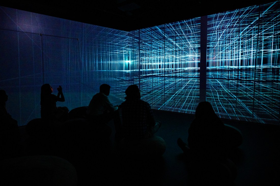 People sit in a dark room watching a glowing digital grid projection at the Data | Art Symposium immersive installation.