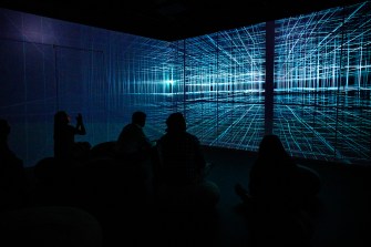 People sit in a dark room watching a glowing digital grid projection at the Data | Art Symposium immersive installation.