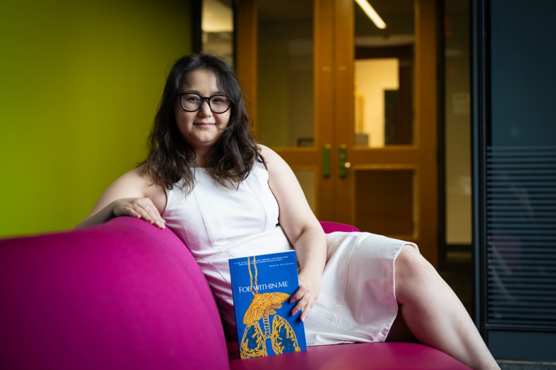 A young woman with glasses sitting on a pink couch, holding a blue book (her memoir) while smiling at the camera.