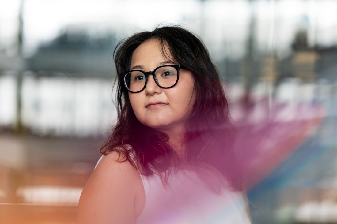 Portrait of Aziza Sattarova wearing glasses with dark hair looking thoughtfully to the side. There is a blurred urban setting in the background.
