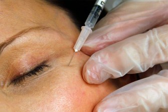 A gloved hand holding a cosmetic needle up to the skin next to a patient's eye, prepared to inject botox into their skin.