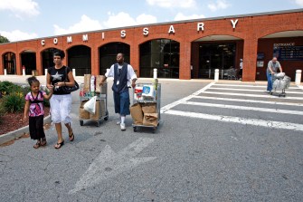 A group of people walking, some moving mobile carts, outside a brick building with "COMMISSARY" spelled out in large white letters across the top