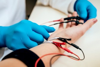 Hands of a medical professional in blue gloves applying electrical stimulation electrodes to a patient's arm during physical therapy treatment.