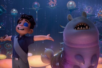Animated character with a bandage over one eye stands joyfully next to a smiling alien creature in a colorful, otherworldly setting from the movie "Elio."