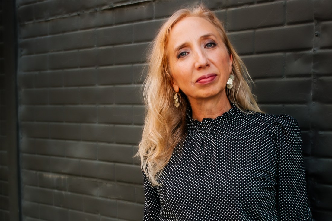 Portrait of Ericka Knudson, standing outside in front of a dark brick wall wearing a polka dot shirt.