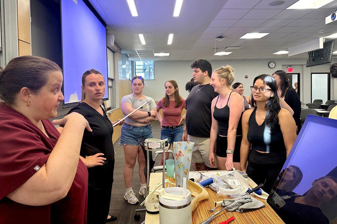 A group of students gathered around a laboratory bench in a classroom setting. One person in a burgundy top appears to be conducting an experiment or demonstration with lab equipment and glassware.