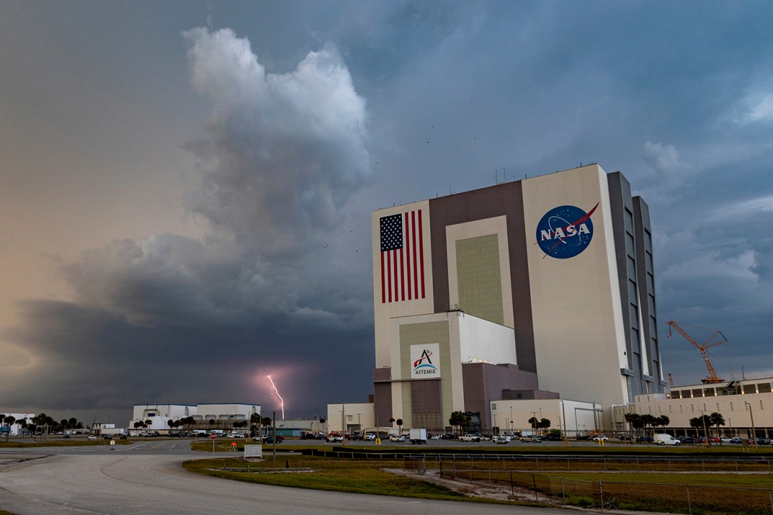 NASA's Vehicle Assembly Building at the Kennedy Space Center with an American flag and NASA logo visible on the exterior, set against a dramatic cloudy sky.