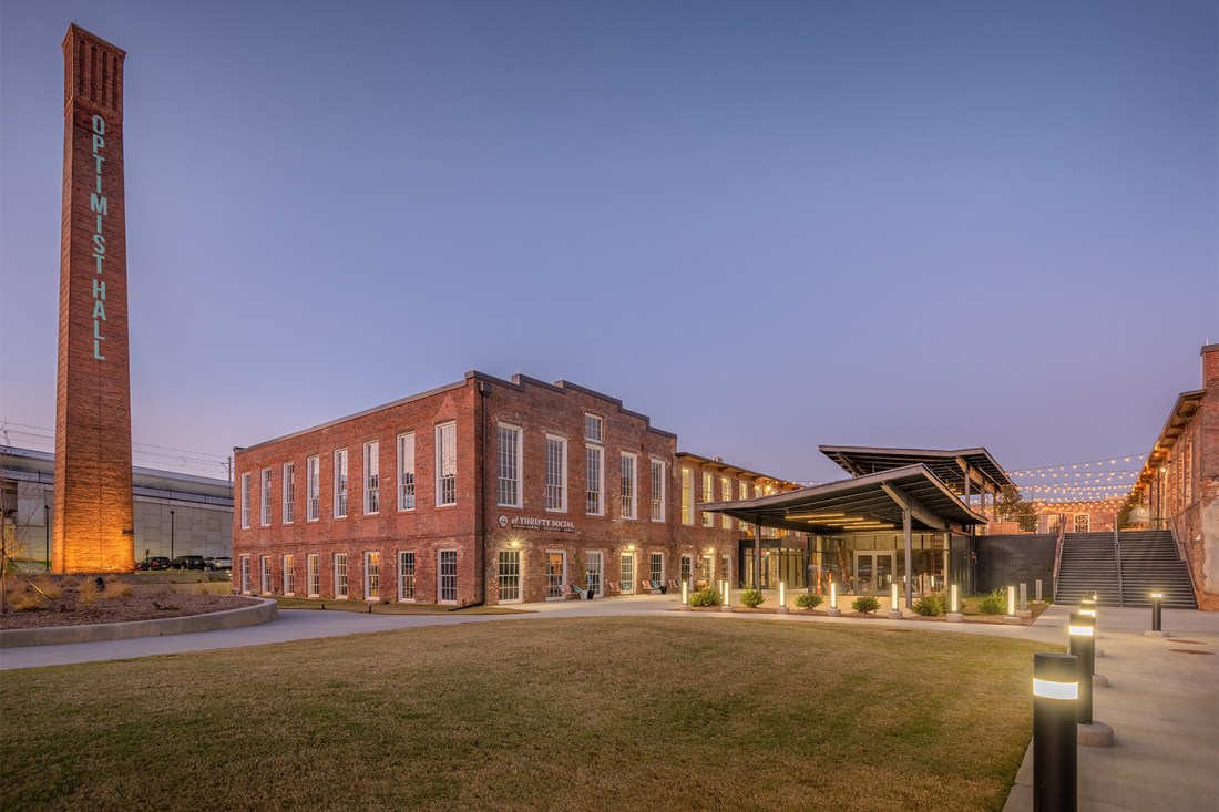 A modern brick building complex photographed at dusk, featuring a  tall cylindrical brick smokestack with 'OPTIMIST' written vertically on it.