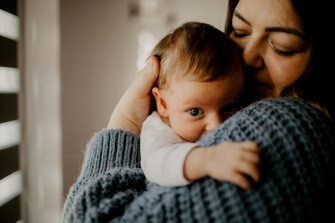 A baby rests against an adult's shoulder in a softly lit room, with the adult holding them close in a comforting embrace.